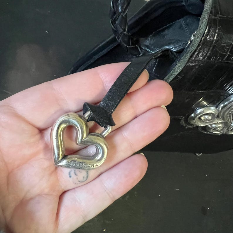 May include: A black leather handbag featuring a textured crocodile-embossed finish and a braided handle. A silver-toned metal heart-shaped charm is attached to the bag by a thin black leather strap, with the brand name Brighton engraved on the charm.
