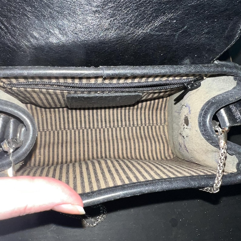 May include: A black handbag with a structured frame and a top flap closure. The interior is lined with a beige fabric featuring thin vertical black stripes. A horizontal zippered pocket is integrated into the back wall of the bag's main compartment.