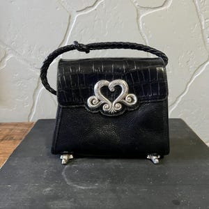 May include: A black handbag with a crocodile-embossed flap and a braided handle. The front features a prominent silver-toned heart-shaped metal ornament. The bag is supported by four small silver-toned feet at the base, providing a stable structure.