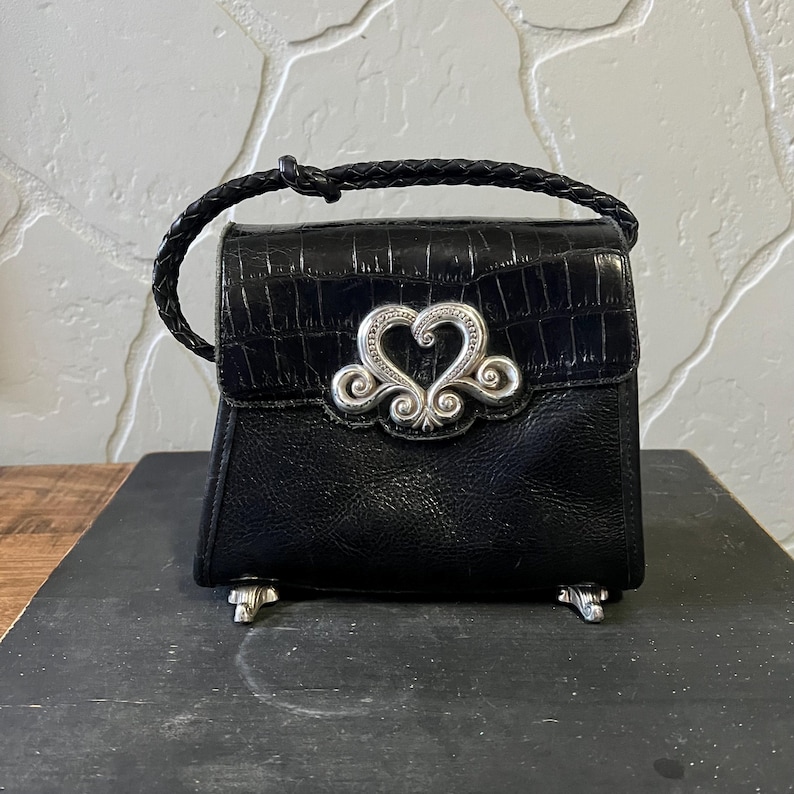 May include: A black handbag with a crocodile-embossed flap and a braided handle. The front features a prominent silver-toned heart-shaped metal ornament. The bag is supported by four small silver-toned feet at the base, providing a stable structure.