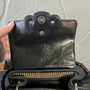 May include: A black leather handbag featuring a scalloped flap closure with a silver-toned magnetic snap button. The interior is lined with a beige and black striped fabric, and the bag includes a braided leather handle and a silver chain strap.