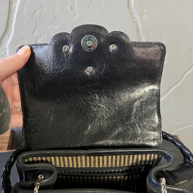 May include: A black leather handbag featuring a scalloped flap closure with a silver-toned magnetic snap button. The interior is lined with a beige and black striped fabric, and the bag includes a braided leather handle and a silver chain strap.