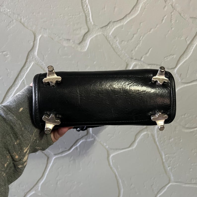 May include: The bottom of a black handbag featuring four silver-toned metal feet. Each foot has a distinct four-pronged, cross-like shape, providing stability and protection for the base of the bag when it is placed on a flat surface.