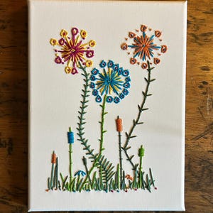 May include: Embroidered floral artwork on a white canvas. The piece features three large, colorful flower blooms in shades of pink, orange, and blue, with green stems and grass. The artwork is mounted on a wooden surface.
