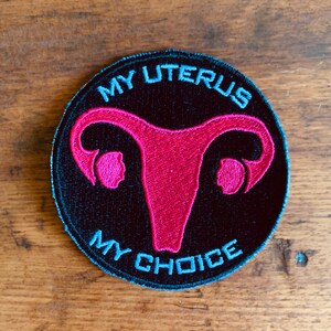 May include: A black and blue embroidered patch with a pink uterus and the text "MY UTERUS MY CHOICE".