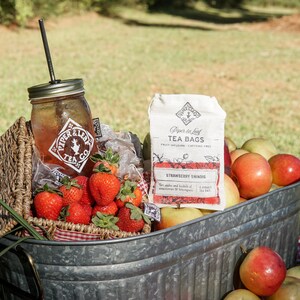 May include: A picnic basket filled with fresh strawberries, a mason jar with a straw, and a bag of "Strawberry Shindig" tea bags. The basket is set in a galvanized metal tub with apples and pears.