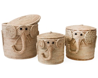 May include: Three woven baskets with elephant faces, the largest basket has its lid open. The baskets are made of natural materials and have a light brown color.