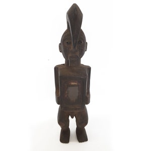 May include: A dark brown wooden sculpture of a person with a tall, pointed headdress. The sculpture has a rectangular shape and a carved face with a serious expression.