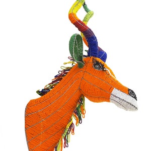 May include: A beaded animal head sculpture of an antelope with long, curled horns. The sculpture is made with colorful beads in shades of orange, green, blue, yellow, and red. The sculpture is a traditional African art form.