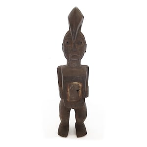 May include: A dark brown wooden sculpture of a person with a pointed head and a carved face. The sculpture is standing with its arms at its sides.