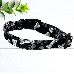 A black and white dog collar with a pattern of bone-shaped paw prints and the word "BONE" printed on it, placed on a white wooden surface with a green plant in the background.