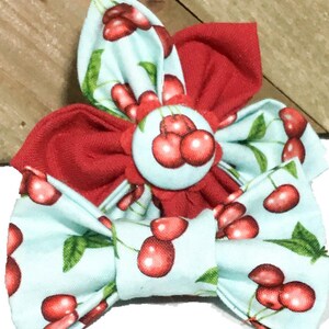 May include: A fabric accessory featuring a light blue bow tie and a flower-shaped ornament, both decorated with a red cherry and green leaf pattern. The center of the flower is a circular button covered in the same light blue cherry fabric.