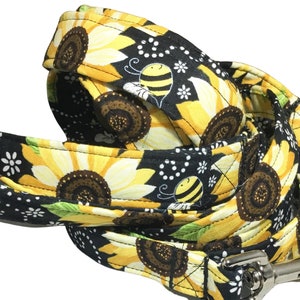 May include: A black and yellow dog leash with a sunflower and bee pattern. The leash has a gold metal clip.
