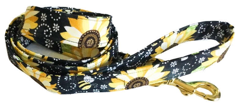 May include: A black and yellow dog leash with a sunflower and bee pattern. The leash has a gold metal clip.