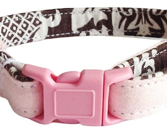 May include: A pet collar featuring a brown and white patterned fabric strap with a light pink plastic side-release buckle. The collar includes a silver-toned metal D-ring and a small silver-toned bell attached to the side of the fabric band.