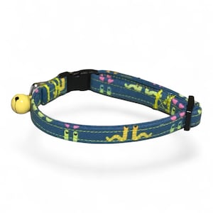 May include: A blue pet collar featuring a repeating pattern of small yellow and pink worm-like figures. The collar includes a black plastic side-release buckle, a black plastic slider, and a small, round, pale yellow metal bell attached to a ring.