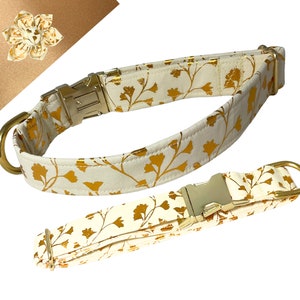 May include: A set of two dog collars with a white fabric base and a gold floral pattern. The collars have gold metal buckles and D-rings.