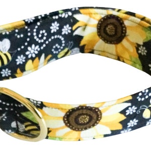May include: A black dog collar with a sunflower and bee pattern. The collar is made of fabric and has a gold metal buckle and D-ring.