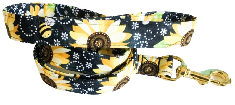 May include: A black dog leash with a sunflower and bee pattern. The leash has a gold metal clip.