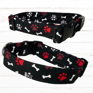May include: Two black dog collars with a red paw print and bone pattern. The collars are made of fabric and have a black buckle.