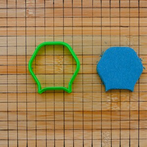 May include: A green plastic cookie cutter in the shape of a hexagon with rounded sides, next to a small blue felt hexagon. The objects are on a wooden cutting board with a grid pattern.