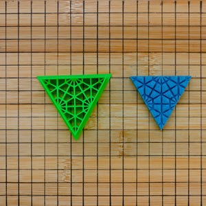 May include: Two geometric triangle shapes, one green and one blue, with a patterned design, are placed on a wooden surface with a grid pattern.
