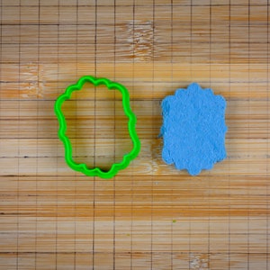 May include: A green plastic cookie cutter shaped like a scalloped oval, next to a blue fondant cookie in the same shape.