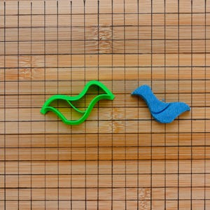 Puede incluir: Un cortador de galletas verde con forma de pájaro y un pájaro azul de fieltro se muestran sobre una tabla de cortar de madera con un patrón de cuadrícula. El cortador de galletas es de color verde brillante, mientras que el pájaro de fieltro es azul liso.