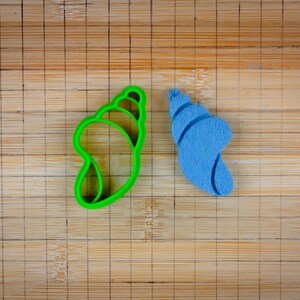 Pode incluir: Dois cortadores de biscoitos em forma de concha, um verde e outro azul, em uma tábua de cortar de bambu.