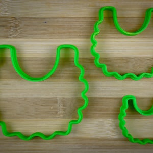 K&ouml;nnte beinhalten: Zwei gr&uuml;ne Plastik-Ausstecher in Form einer L&auml;tzchen, einer gr&ouml;&szlig;er als der andere, auf einer Holzoberfl&auml;che.
