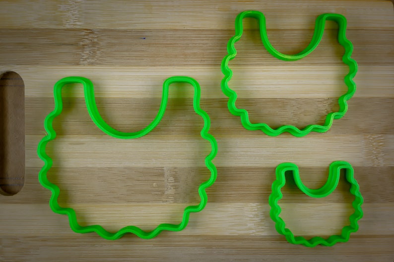 K&ouml;nnte beinhalten: Zwei gr&uuml;ne Plastik-Ausstecher in Form einer L&auml;tzchen, einer gr&ouml;&szlig;er als der andere, auf einer Holzoberfl&auml;che.