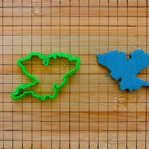 May include: A green plastic cookie cutter and a blue felt cutout, both in the shape of a geographical region, rest on a bamboo cutting board. The cookie cutter is a bright, vibrant green, while the felt cutout is a solid, medium blue.