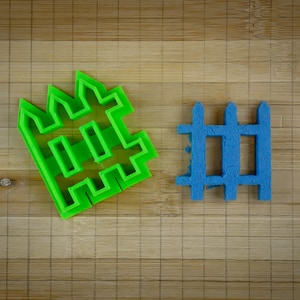 May include: Two plastic cookie cutters shaped like fences. The larger cutter is green and has a more elaborate design. The smaller cutter is blue and has a simpler design.