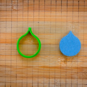 Può includere: Un tagliapasta di plastica verde e una goccia di pioggia di feltro blu su un tagliere di legno.