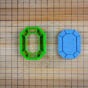 May include: A green plastic gemstone-shaped cookie cutter next to a blue, gemstone-shaped sponge. The objects are on a bamboo cutting board with a grid pattern.