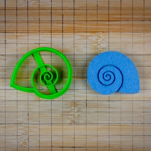 May include: Two shell-shaped cookie cutters, one green and one blue, on a bamboo cutting board.