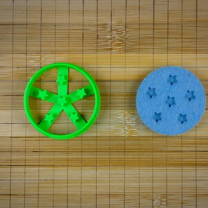 Pode incluir: Dois cortadores de biscoitos sobre uma superfície de madeira. Um é verde e tem a forma de uma estrela de cinco pontas. O outro é azul e tem forma circular com cinco recortes em forma de estrela.