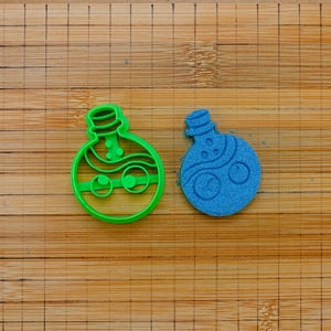 Peut inclure: Un emporte-pièce en plastique vert vif en forme de flacon de potion à côté d'une découpe de pâte ou d'un biscuit bleu en forme de flacon. Les deux objets présentent une base ronde, un goulot étroit avec un bouchon et un motif ondulé.