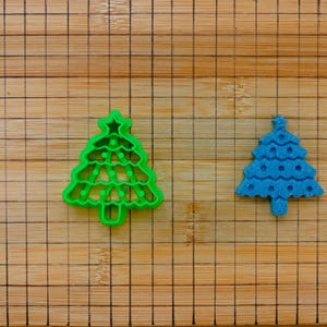 Könnte beinhalten: Zwei Ausstecher in Form eines Weihnachtsbaums. Einer ist leuchtend grün, der andere blau. Der grüne Ausstecher hat einen Stern oben. Beide liegen auf einem Holzschneidebrett.