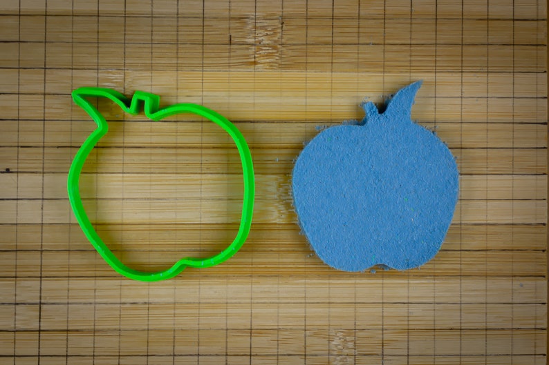 Peut inclure: Un emporte-pi&egrave;ce en forme de pomme en plastique vert &agrave; c&ocirc;t&eacute; d'une pomme en feutre bleu.