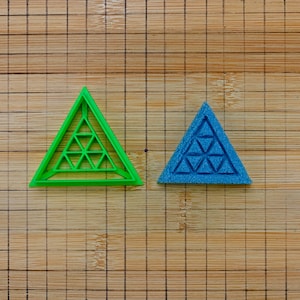 Può includere: Due formine triangolari, una verde e una blu, sono appoggiate su un tagliere di legno. Ogni formina ha un design geometrico all'interno della forma triangolare. La tavola ha un motivo a griglia.