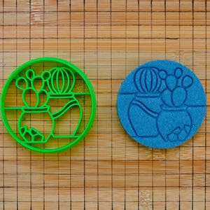 Könnte beinhalten: Ein grüner Kaktus-Ausstecher aus Kunststoff neben einem blauen Abdruck desselben Designs. Der Ausstecher ist ein Kreis mit einem Kaktus- und Topfdesign. Der blaue Abdruck befindet sich auf einem hellbraunen Bambus-Schneidebrett.