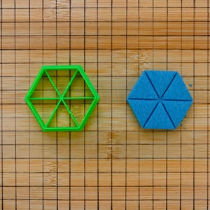 May include: A green plastic hexagon-shaped cookie cutter with internal lines, next to a blue hexagon-shaped object with triangular segments. Both are on a wooden cutting board with a grid pattern.