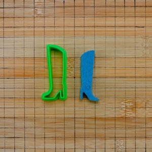 May include: Two boot-shaped cookie cutters. One is green, the other is blue. The blue boot has a textured pattern. Both cutters are on a wooden cutting board.