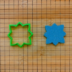 Könnte beinhalten: Ein grüner Ausstecher in Sternform neben einer blauen Sternform aus Filz. Beide liegen auf einem Holzschneidebrett mit einem Rastermuster.