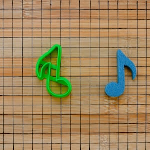 May include: Two music note cookie cutters, one green and one blue, on a brown bamboo cutting board.