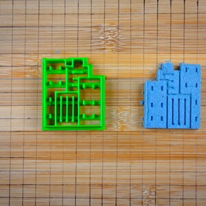 以下が含まれることがあります： 木製のまな板の上に置かれた2つのクッキーカッター。左側のクッキーカッターは緑色で、建物のような形をしています。右側のクッキーカッターは青色で、建物のような形をしています。