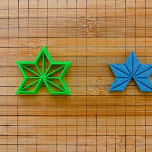 May include: Two star-shaped cookie cutters. One is bright green and the other is blue. The green cutter has a geometric design inside. The blue cutter has a folded paper appearance. Both are on a bamboo cutting board.