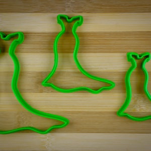May include: Three green plastic cookie cutters shaped like dresses. The cutters are on a wooden surface.