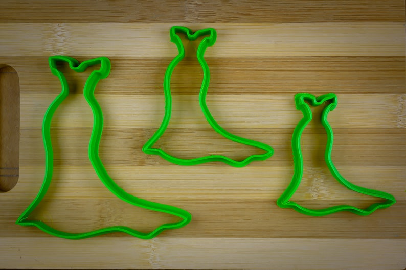 May include: Three green plastic cookie cutters shaped like dresses. The cutters are on a wooden surface.